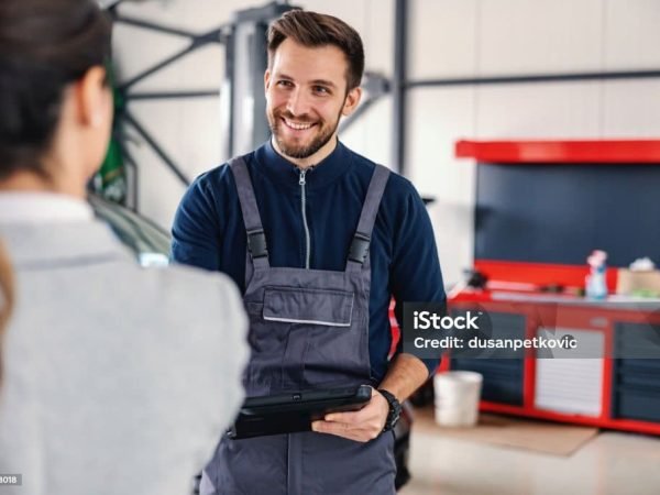 Smiling friendly car seller talking to a mechanic and showing okay hand gesture while standing in car salon.